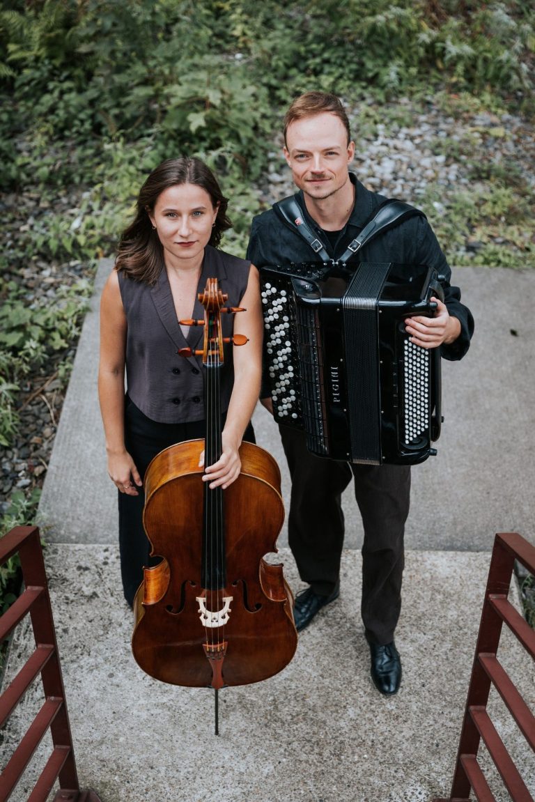 Zwei Musiker, einer mit Akkordeon und die andere mit Cello, stehen vor einer Backsteinmauer.