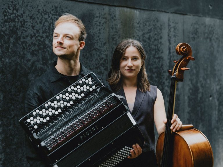 Zwei Musiker in urbaner Umgebung; einer mit Akkordeon, die andere mit Violoncello.